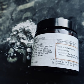 A close-up view of a labeled jar placed on a dark textured surface with a pile of powder behind it. The jar features text about the product, specifying 'Gunpowder Fragrance' and other details such as the hold, finish, and ingredients.