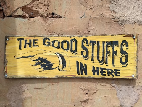 A vintage yellow wooden sign with black lettering and an illustration of a pointing hand. The text reads 'THE GOOD STUFFS IN HERE.' The sign is mounted on a rough, textured wall with visible cracks and a weathered appearance.