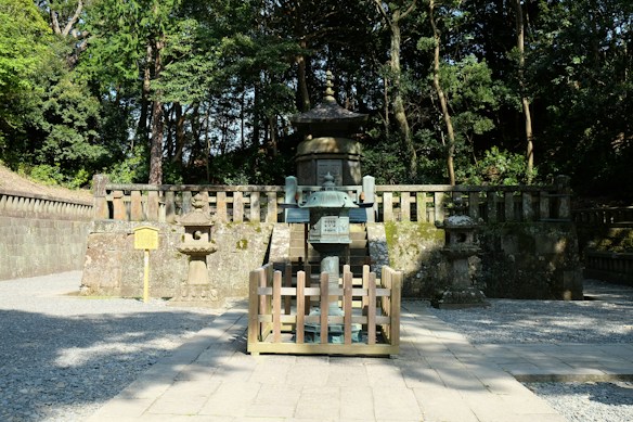 A traditional Japanese stone lantern and shrine structure set within a dense, lush green forest. The stonework shows weathering and moss growth, indicating age. Gravel paths and a wooden fence add to the tranquil and historical atmosphere.