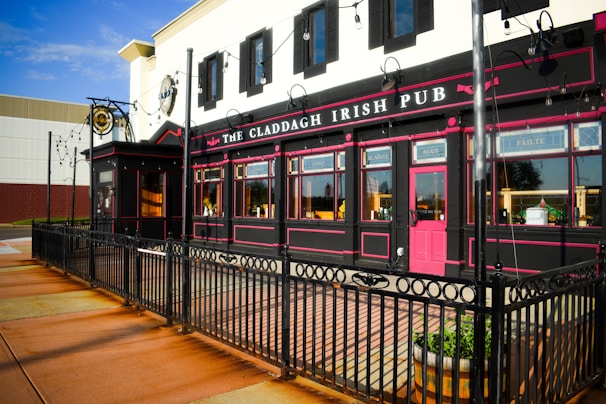 An Irish pub with black and pink exterior details, featuring outdoor alley-like seating enclosed by a black iron fence. The building is adorned with string lights and wooden barrel planters. It has large windows with decorative signage and a distinctive red door.