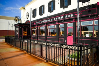 An Irish pub with black and pink exterior details, featuring outdoor alley-like seating enclosed by a black iron fence. The building is adorned with string lights and wooden barrel planters. It has large windows with decorative signage and a distinctive red door.