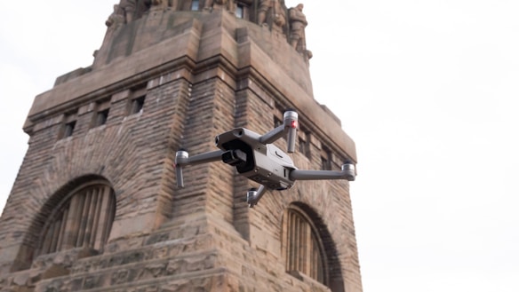 A modern drone hovers in front of an old, intricately designed stone monument with arched windows and statues at the top. The drone appears metallic and sleek, contrasting with the historic structure.