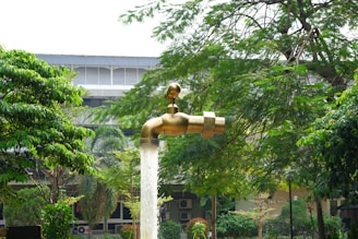 A large, metallic, floating tap appears to be suspended in mid-air with water flowing from it into a pool below. The illusion is set in a garden or park-like setting with lush green trees and a building in the background.