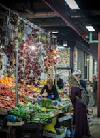A bustling indoor market scene with a vibrant display of fresh fruits and vegetables. A woman working behind the stall is arranging produce while two customers examine the selection. Above the stall, dried peppers hang in clusters, adding to the lively atmosphere.