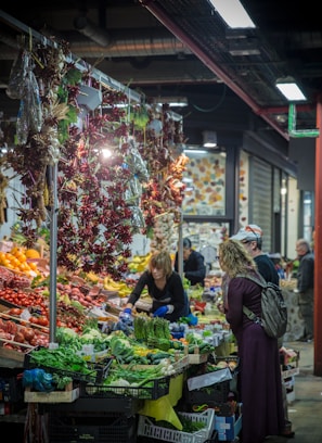 A bustling indoor market scene with a vibrant display of fresh fruits and vegetables. A woman working behind the stall is arranging produce while two customers examine the selection. Above the stall, dried peppers hang in clusters, adding to the lively atmosphere.