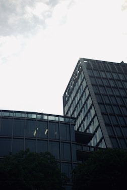 A modern high-rise office building with reflective glass windows set against a cloudy sky. The architecture is sleek and contemporary, with numerous floors and a geometric design. In the foreground, some trees are visible, creating a contrast between nature and the urban environment.