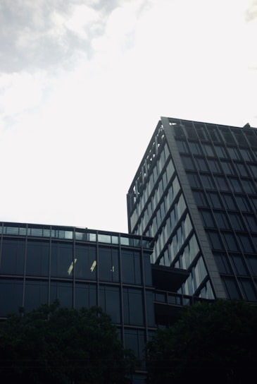 A modern high-rise office building with reflective glass windows set against a cloudy sky. The architecture is sleek and contemporary, with numerous floors and a geometric design. In the foreground, some trees are visible, creating a contrast between nature and the urban environment.