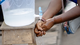 A person washing hands under a small tap attached to a transparent container placed on a wooden stand. Water is flowing onto the hands, with droplets visible. The person is wearing bracelets, and there is a bag hanging from the shoulder.
