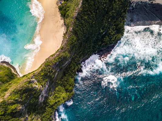 Aerial view of a picturesque beach with turquoise water meeting golden sand, bordered by lush green cliffs. Waves crash against the rugged shoreline, creating a dramatic contrast with the serene beach area. The natural landscape is vibrant and unspoiled, evoking a sense of tranquility and isolation.