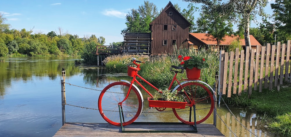 red city bike parked beside brown wooden house near body of water during daytime