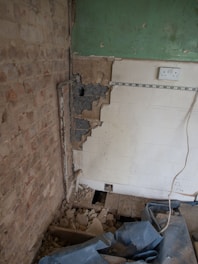 A partially demolished room with exposed brick walls and broken plaster. The floor is covered in debris and rubble. Visible are sections of green painted wall, white tiles, an electrical outlet, and an uncovered floor area. There is also a length of cable extending across the scene.
