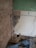 A partially demolished room with exposed brick walls and broken plaster. The floor is covered in debris and rubble. Visible are sections of green painted wall, white tiles, an electrical outlet, and an uncovered floor area. There is also a length of cable extending across the scene.