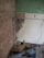 A partially demolished room with exposed brick walls and broken plaster. The floor is covered in debris and rubble. Visible are sections of green painted wall, white tiles, an electrical outlet, and an uncovered floor area. There is also a length of cable extending across the scene.