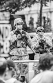 Two soldiers in camouflage uniforms and helmets stand with rifles at a public event. They wear sunglasses and appear serious and alert. The background shows blurred outlines of a building and greenery.