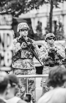 Two soldiers in camouflage uniforms and helmets stand with rifles at a public event. They wear sunglasses and appear serious and alert. The background shows blurred outlines of a building and greenery.