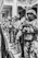 A line of uniformed, armed personnel stand in a row on the street near a building, holding weapons and wearing helmets and protective gear.
