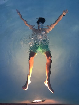 A person is floating in a swimming pool, viewed from underneath. The person's arms are stretched out to the sides, and they are wearing swim trunks. The pool light illuminates their legs, creating interesting light effects in the water.