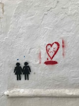 Graffiti painted on a white textured wall depicts two figures holding hands in black, each with a small blue dot above their heads. To the right is a red heart with a sad face inside, appearing to cry with drops at the bottom.