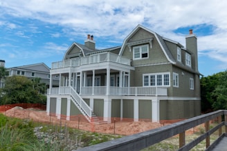 A two-story house with a large porch and multiple gables. The exterior features a muted green wooden finish with white trim and railings. There are two chimneys, and a stairway leads to the raised front porch. Surrounding the house are tall grasses and some greenery, with an orange safety fence indicating construction or landscaping work nearby.