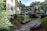 A well-maintained landscaped courtyard features a brick pathway lined with flowering plants and bushes. Palm trees and manicured shrubs enhance the tropical setting, with houses and parked cars visible in the background. The scene is bathed in natural light, giving a bright and inviting atmosphere.