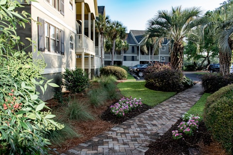 A well-maintained landscaped courtyard features a brick pathway lined with flowering plants and bushes. Palm trees and manicured shrubs enhance the tropical setting, with houses and parked cars visible in the background. The scene is bathed in natural light, giving a bright and inviting atmosphere.