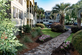 A well-maintained landscaped courtyard features a brick pathway lined with flowering plants and bushes. Palm trees and manicured shrubs enhance the tropical setting, with houses and parked cars visible in the background. The scene is bathed in natural light, giving a bright and inviting atmosphere.