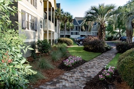 A well-maintained landscaped courtyard features a brick pathway lined with flowering plants and bushes. Palm trees and manicured shrubs enhance the tropical setting, with houses and parked cars visible in the background. The scene is bathed in natural light, giving a bright and inviting atmosphere.