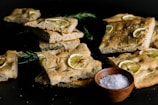 Slices of focaccia bread topped with thin slices of lemon and sprigs of rosemary are arranged on a dark surface. A wooden bowl filled with coarse sea salt is also present, enhancing the rustic and artisanal presentation of the bread.