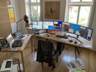 An office space with a large wooden desk featuring an extensive multi-monitor setup. The monitors display various charts and graphs, suggesting stock or data analysis. There is a laptop and additional electronics on the desk, along with some scattered papers, a lamp, and a few drinking glasses. A chair with a jacket on it is positioned in front of the desk. The room is bright, with light streaming in through large windows, and a piece of art hangs on the wall.