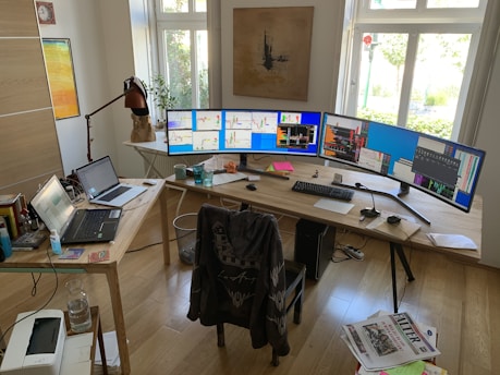 An office space with a large wooden desk featuring an extensive multi-monitor setup. The monitors display various charts and graphs, suggesting stock or data analysis. There is a laptop and additional electronics on the desk, along with some scattered papers, a lamp, and a few drinking glasses. A chair with a jacket on it is positioned in front of the desk. The room is bright, with light streaming in through large windows, and a piece of art hangs on the wall.