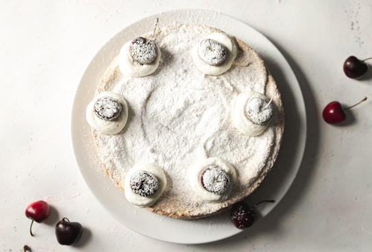 A round cake delicately dusted with powdered sugar, topped with perfectly whipped cream and fresh cherries. The cake sits on a white plate with additional cherries scattered around on the surface.