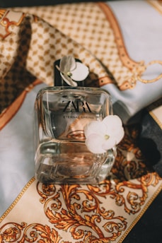 A perfume bottle labeled 'ZARA ETERNAL SPRING' is positioned on ornate fabric with intricate gold and brown patterns. A delicate white flower is attached to the corner of the bottle, adding a touch of elegance.