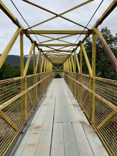 A yellow metal bridge with a series of crossbeams and railings spans across a natural setting. The bridge has a grated floor and is surrounded by lush green trees and distant hills under a cloudy sky.