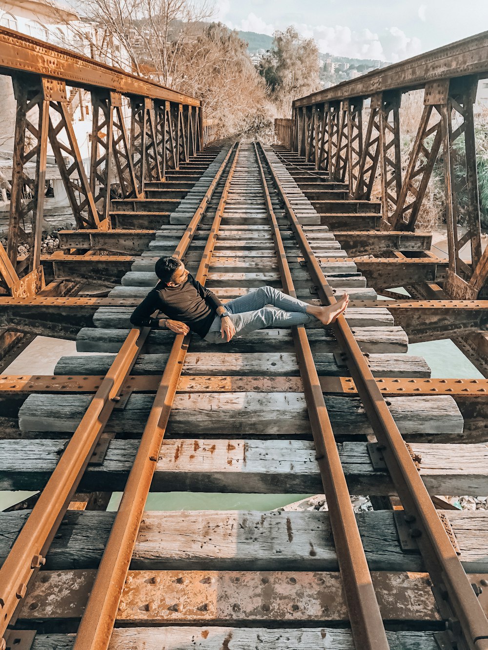 man in black jacket and blue denim jeans sitting on brown wooden bridge during daytime