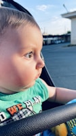 A baby with short hair and big eyes gazes attentively to the right while seated outside in a stroller. The baby wears a green shirt with colorful text and appears to be enjoying an evening outdoors, as the soft lighting suggests a sunset. The background shows an out-of-focus urban scene with buildings and the sky.