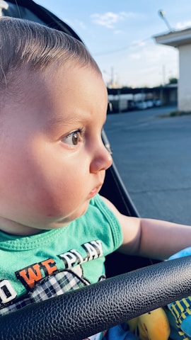 A baby with short hair and big eyes gazes attentively to the right while seated outside in a stroller. The baby wears a green shirt with colorful text and appears to be enjoying an evening outdoors, as the soft lighting suggests a sunset. The background shows an out-of-focus urban scene with buildings and the sky.