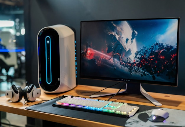 A sleek gaming setup featuring a large computer monitor displaying a colorful, cosmic scene with planets and space debris. The desktop tower is uniquely designed with a vertical blue light strip. A gaming keyboard with rainbow-colored backlighting and a gaming mouse are placed on a dark-colored mat. A pair of headphones rests on the desk alongside the tower, contributing to the modern aesthetic of the workspace.