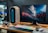 A sleek gaming setup featuring a large computer monitor displaying a colorful, cosmic scene with planets and space debris. The desktop tower is uniquely designed with a vertical blue light strip. A gaming keyboard with rainbow-colored backlighting and a gaming mouse are placed on a dark-colored mat. A pair of headphones rests on the desk alongside the tower, contributing to the modern aesthetic of the workspace.