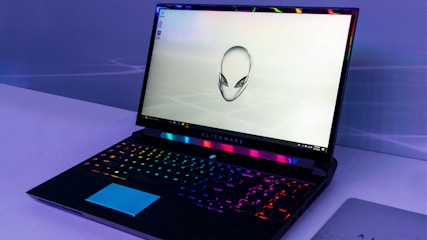 A sleek gaming laptop with a vibrant backlit keyboard and an iconic alien head logo displayed on the screen. The laptop sits on a minimalistic surface, and the lighting around the device creates a futuristic ambiance.