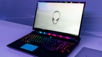A sleek gaming laptop with a vibrant backlit keyboard and an iconic alien head logo displayed on the screen. The laptop sits on a minimalistic surface, and the lighting around the device creates a futuristic ambiance.