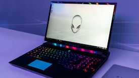 A sleek gaming laptop with a vibrant backlit keyboard and an iconic alien head logo displayed on the screen. The laptop sits on a minimalistic surface, and the lighting around the device creates a futuristic ambiance.