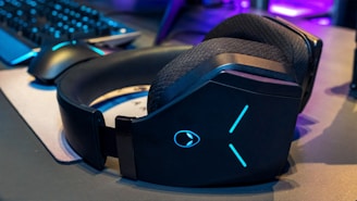 A pair of large black over-ear headphones with glowing blue details sits on a gaming desk. Nearby, a gaming mouse and keyboard are visible, with a soft light reflecting off their surfaces. The atmosphere suggests a gaming setup with a mix of technology and ambient lighting.