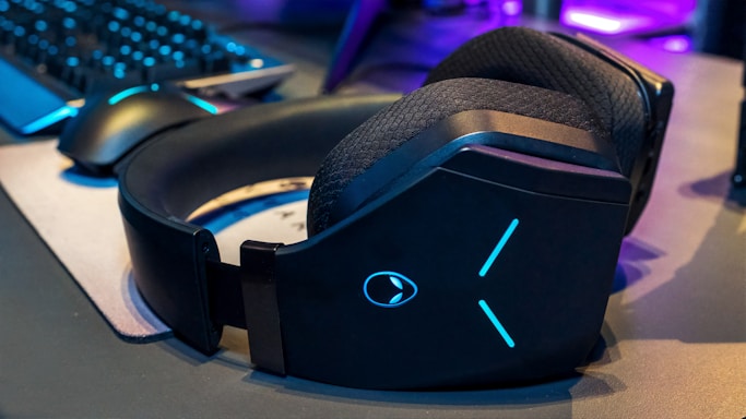 A pair of large black over-ear headphones with glowing blue details sits on a gaming desk. Nearby, a gaming mouse and keyboard are visible, with a soft light reflecting off their surfaces. The atmosphere suggests a gaming setup with a mix of technology and ambient lighting.