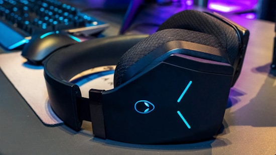 A pair of large black over-ear headphones with glowing blue details sits on a gaming desk. Nearby, a gaming mouse and keyboard are visible, with a soft light reflecting off their surfaces. The atmosphere suggests a gaming setup with a mix of technology and ambient lighting.
