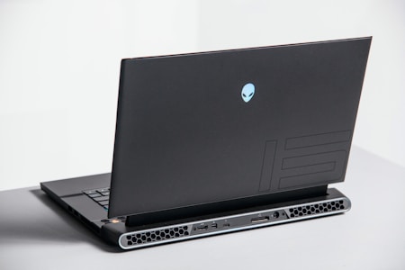 A sleek, dark laptop with a futuristic design, showcasing a blue alienware logo on its back cover. The laptop has a robust hinge, visible ventilation slots, and several ports at the back. It's placed on a light gray surface, with part of the slightly open keyboard in view.