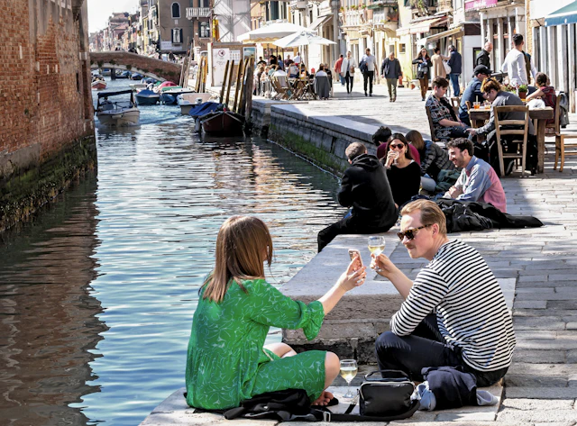 A couple toast glasses of wine while sitting on canal-side steps along a Cannaregio canal lined with cafes.