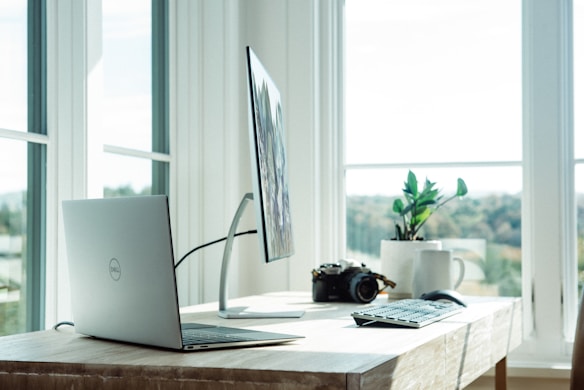A workspace setup near a large window with a laptop, a monitor on a stand, and a keyboard and mouse on a wooden desk. A camera and a mug are placed beside a potted plant. Natural light fills the room, creating a bright and serene atmosphere.