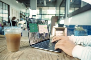 A person types on a laptop in a modern, well-lit cafe. The wooden table has a cold beverage with a straw beside the laptop. The screen displays a calendar and other applications. The environment includes contemporary decor with plants and blurred background figures.