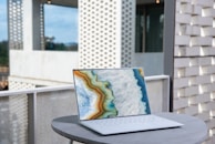 A sleek laptop with a vibrant abstract design on its screen is placed on a round, light grey table. The backdrop includes a modern architectural setting with perforated brick walls and a partially visible glass facade.