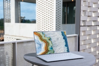 A sleek laptop with a vibrant abstract design on its screen is placed on a round, light grey table. The backdrop includes a modern architectural setting with perforated brick walls and a partially visible glass facade.